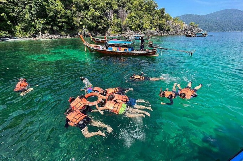 People swimming in clear water with boats and a forested island in the background. 1
