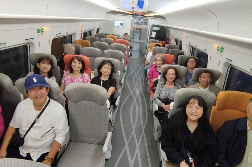 A train interior with passengers seated, overhead luggage compartments, and a patterned floor. 1