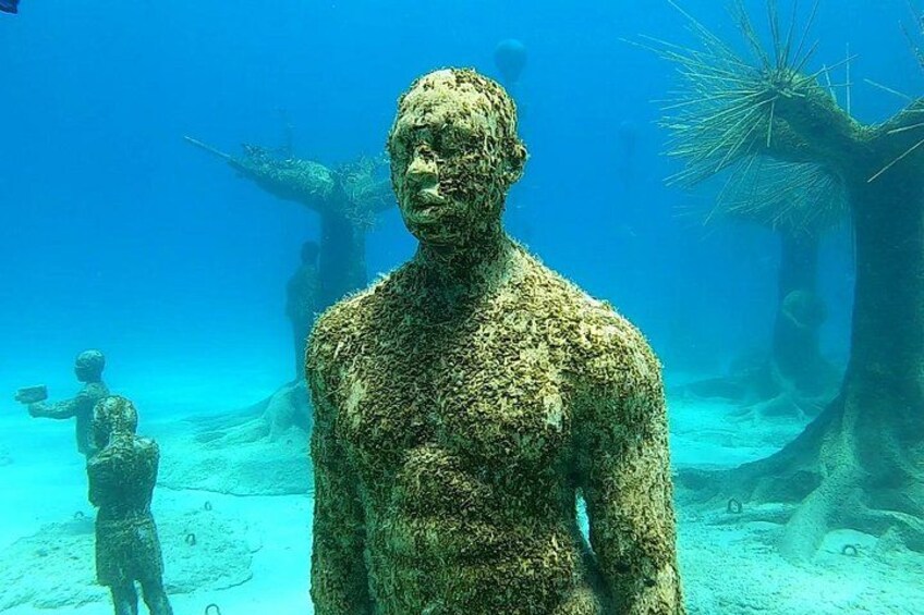 En stor undervands skulptur, der ligner en menneskelig figur, omgivet af mindre statuer og havplanter. 4