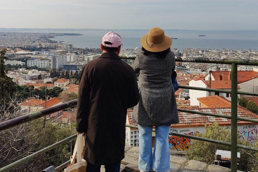 Two people standing on a balcony overlooking a cityscape with buildings and a body of water. 4