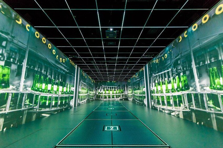 Un espacio industrial largo y estrecho con hileras de botellas de vidrio verde en estanterías, un techo enrejado y un pasillo central. 4