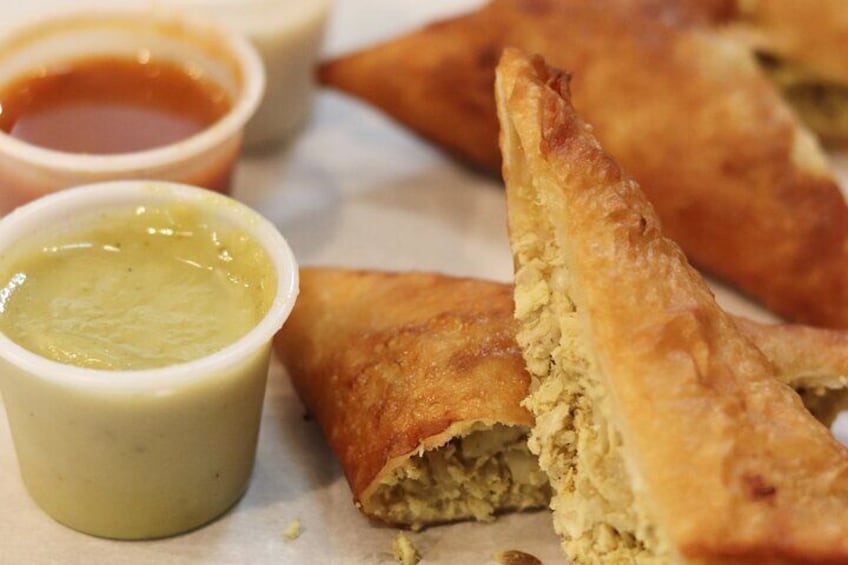 A food item, possibly a pastry, with a crispy exterior and a filling that appears to be a mixture of ingredients. There are two small containers with sauces, one possibly a green chutney and the other a brown sauce. 5