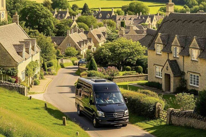 A black van driving down a narrow road in a rural village with stone houses and lush greenery. 1