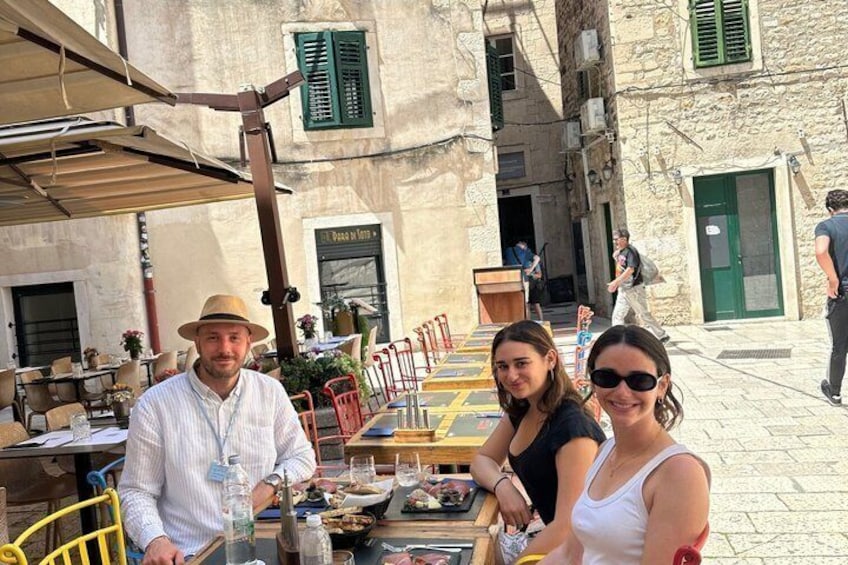 Un groupe de personnes est assis à un café en plein air, autour de tables et de chaises. On aperçoit un bâtiment à la façade en pierre et, au loin, une rue. 2
