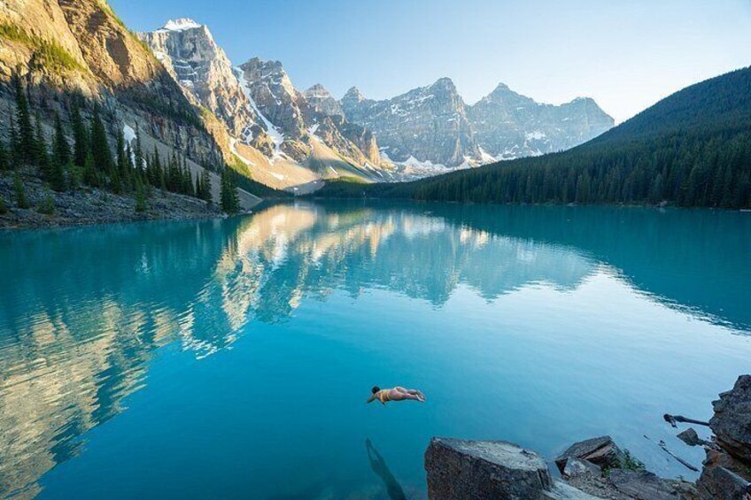 A calm lake with a swimmer, surrounded by mountains and dense forest. 5