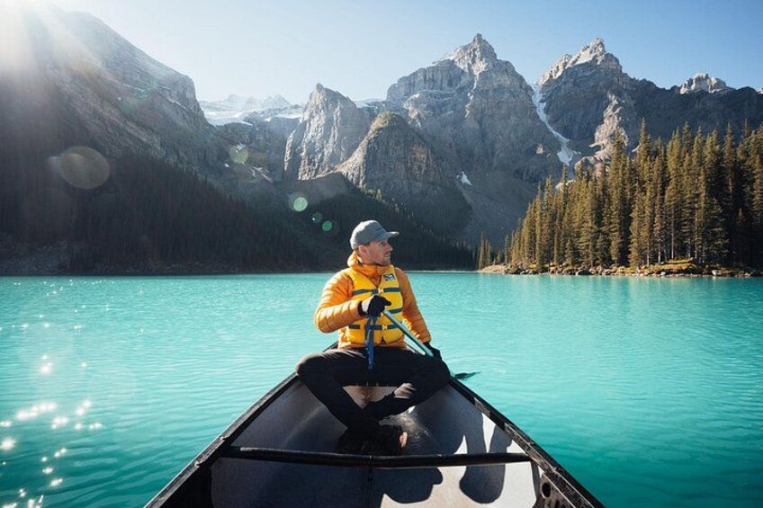 A person in a yellow jacket paddling a canoe on turquoise water with mountains in the background. 2
