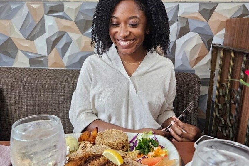 A person sitting at a table with a meal, a glass of water, and a decorative wall in the background. 4