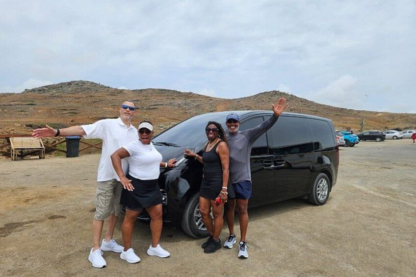 A group of people posing in front of a black van in a parking lot with a hilly landscape in the background. 1