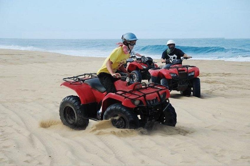 Twee personen rijden op rode quads over een zandstrand, met de oceaan op de achtergrond. 5
