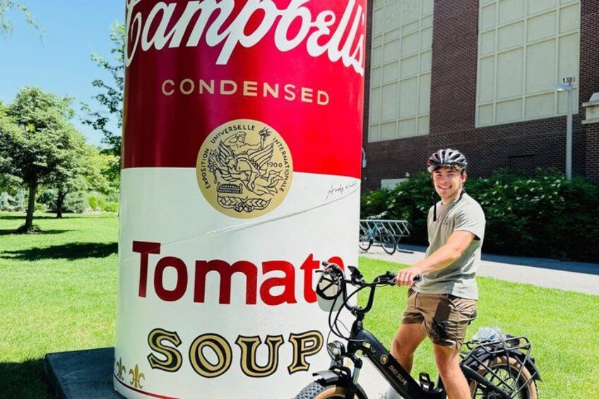 A person on a bicycle next to a large can of Campbell's Tomato Soup. 1