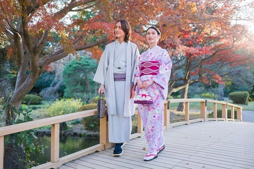 Two people in traditional Japanese attire on a wooden bridge with autumn foliage in the background. 1