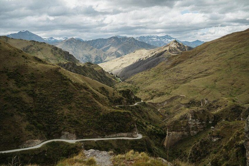 Una carretera sinuosa serpentea por un valle con terreno accidentado y montañas nevadas a lo lejos. 1