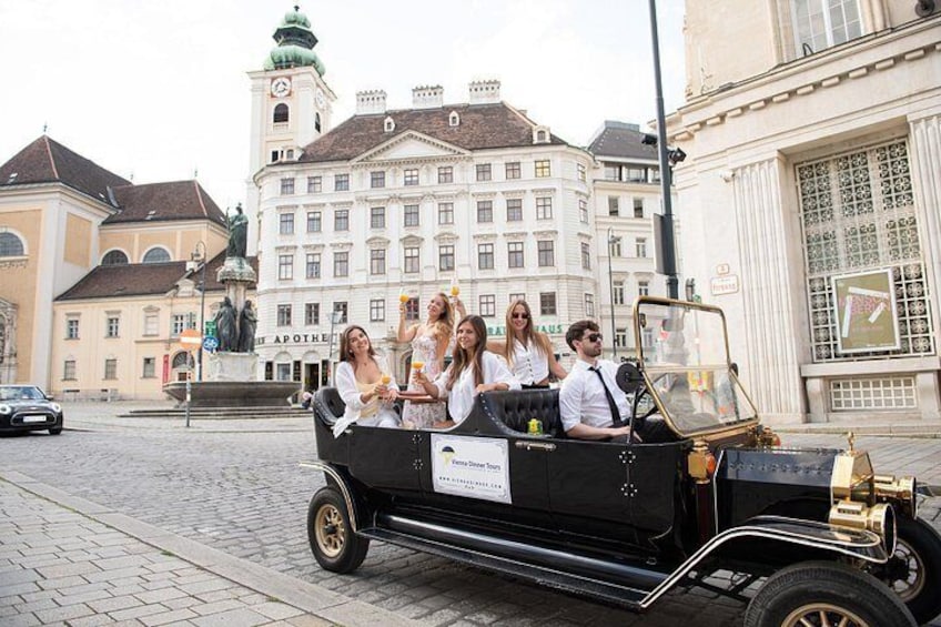 A vintage car with passengers, a historic building, and a clock tower in the background. 2