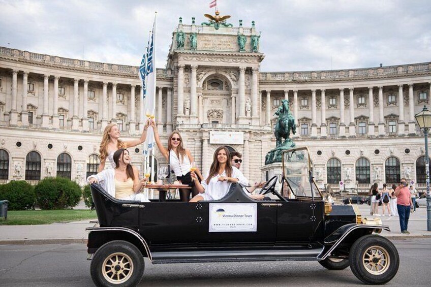 A vintage car with passengers, a building with columns, and a statue in the background. 1