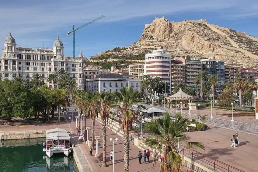 Un paisaje urbano costero con una colina destacada, un edificio blanco con cúpulas, una embarcación atracada en el paseo marítimo y un paseo peatonal con palmeras. 3