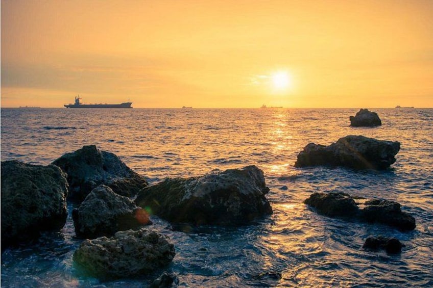 A sunset over the ocean with rocks in the foreground and a ship in the distance. 2