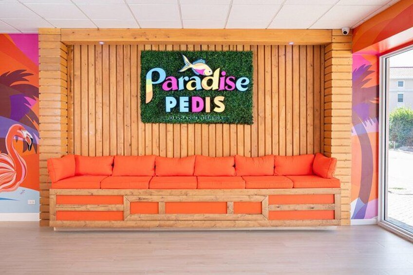 Un banco de madera con cojines anaranjados, una pared de madera con listones verticales y un letrero que dice 'Paradise PEDIS'. 3