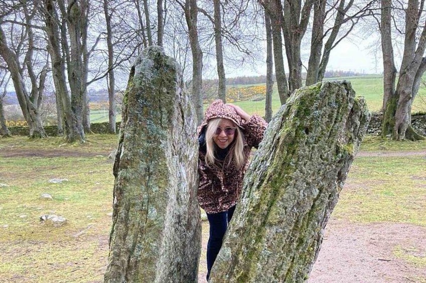 A person standing between two large moss-covered rocks in a natural setting with trees and a grassy field in the background. 4