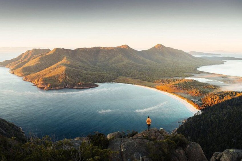 A coastal landscape with a person standing on a cliff overlooking a bay, surrounded by hills and a clear sky. 5