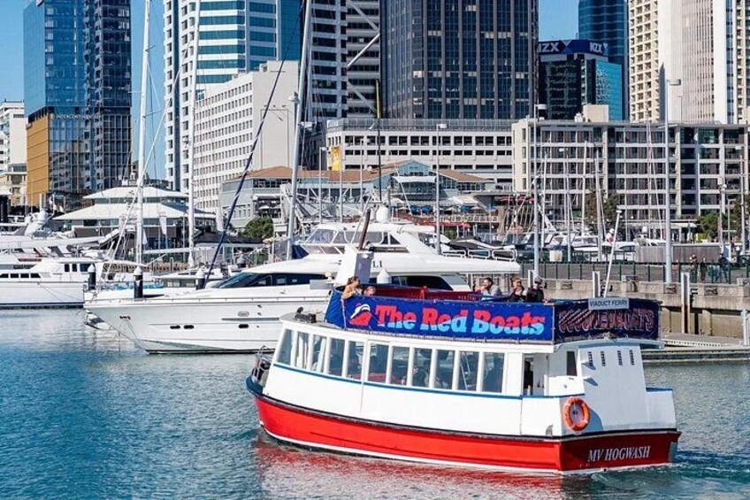 Un bote rojo y blanco llamado 'The Red Boats' con un letrero que dice 'MY HOGWASHI' en el costado, amarrado en un puerto deportivo con otros botes y edificios de la ciudad al fondo. 4