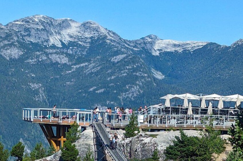 A mountain observation deck with people, overlooking a forested valley and snow-capped mountains. 3
