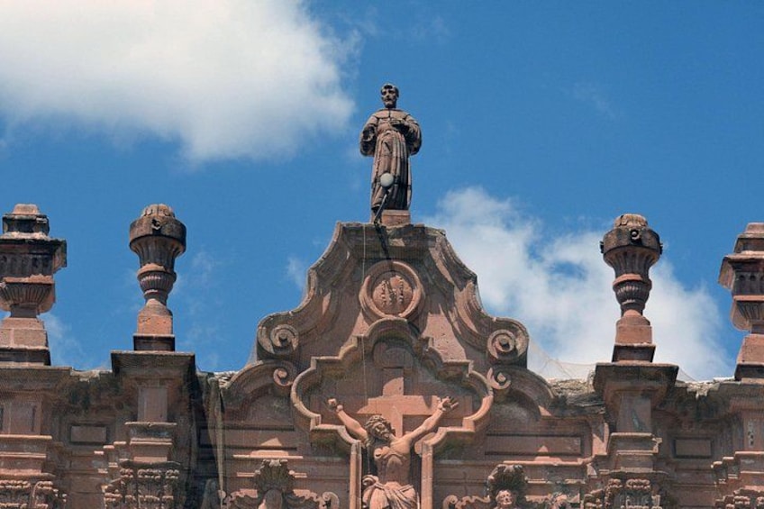 A large stone statue atop a building with intricate carvings and multiple smaller statues around it. 1