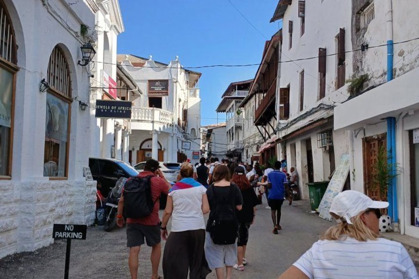 A narrow street lined with white buildings, a 'No Parking' sign, and people walking. 1