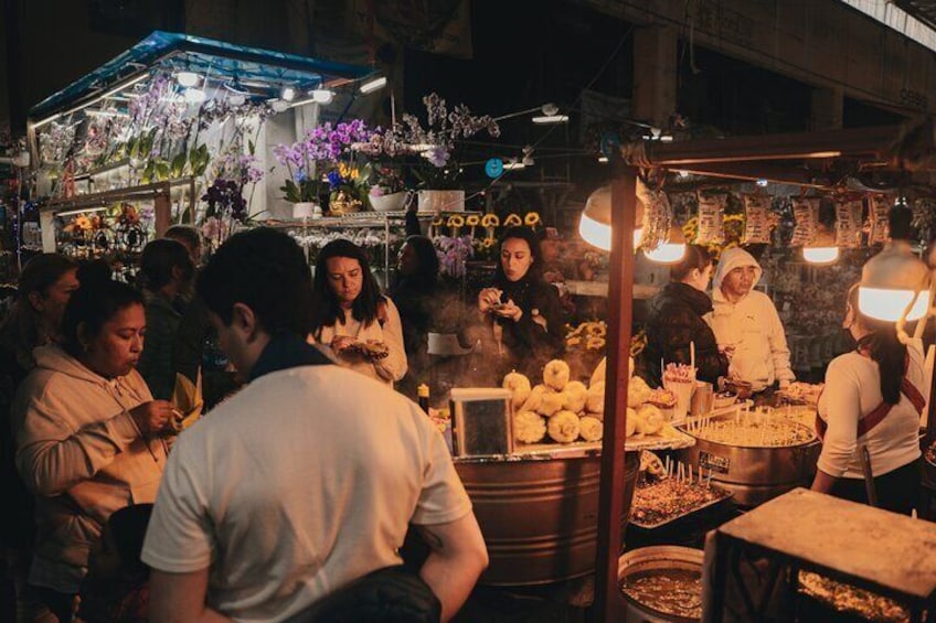 Un mercado de comida callejera concurrido, con gente reunida alrededor de puestos de comida, una olla grande y una variedad de flores y plantas. 1