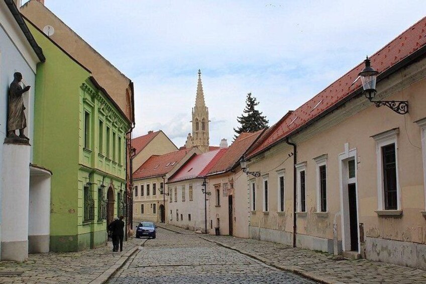 A cobblestone street lined with historic buildings, a church spire in the distance, and a person walking. 4