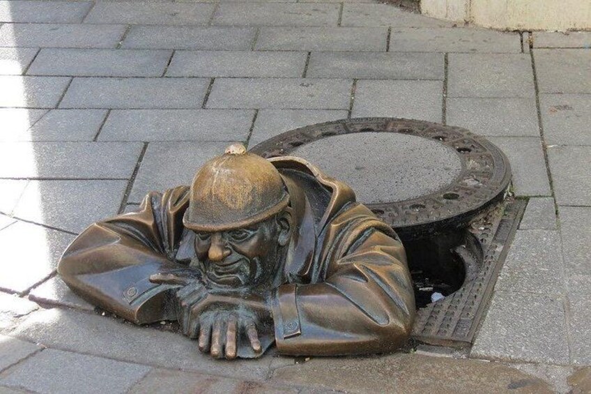 A bronze statue of a man lying down with his head resting on his hand, positioned on a paved surface next to a circular manhole cover. 1