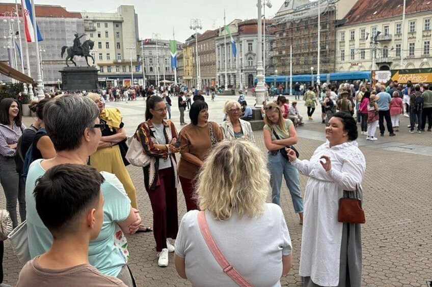 A busy public square with people gathered, a statue, and buildings in the background. 2