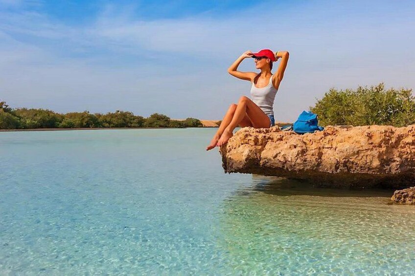 Una persona sentada sobre un tronco junto a una masa de agua azul cristalina, con follaje verde al fondo. 4