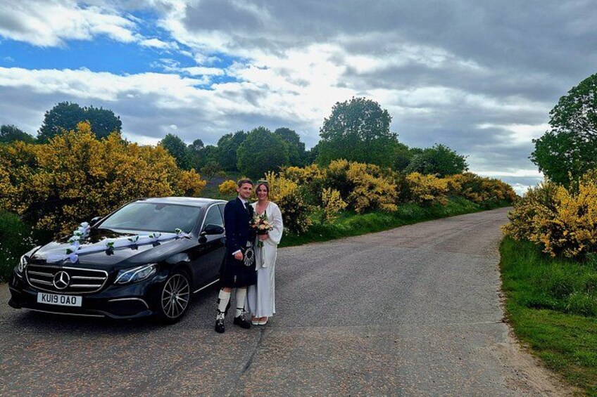 A couple standing next to a black car on a rural road with yellow flowering bushes and trees in the background. 2