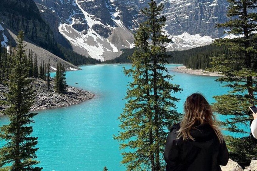 Une personne se tient au bord d’un lac turquoise, entouré de montagnes enneigées et d’arbres à feuilles persistantes. 4