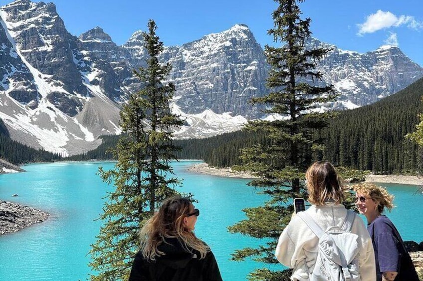 A group of people standing by a turquoise lake with snow-capped mountains in the background. 1