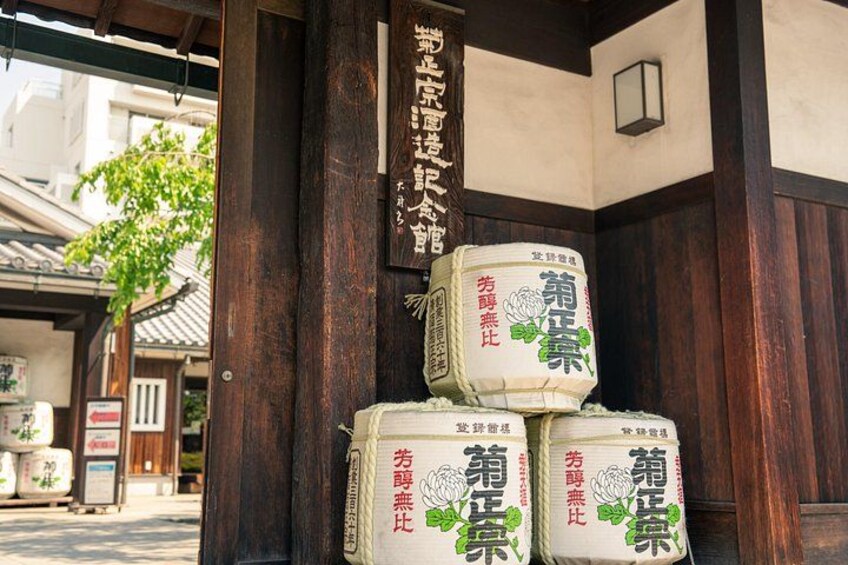 Il s'agit d'un bâtiment japonais traditionnel, avec des poutres en bois et une enseigne. On peut y voir trois grands sacs de sauce soja empilés sur une plateforme en bois. 3