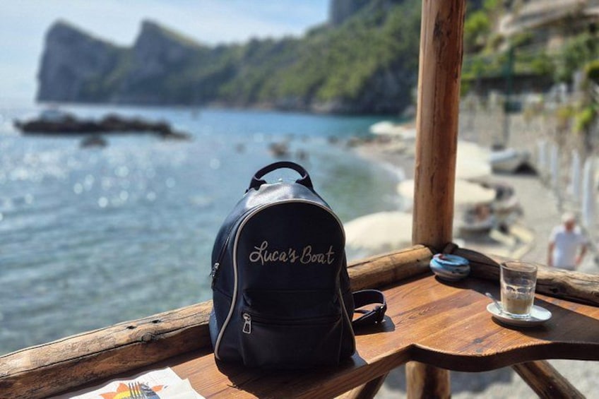 Ein schwarzer Rucksack mit der Aufschrift 'Luca's Boat', ein Holztisch, ein Glas Wasser und ein reizvoller Blick auf einen Strand mit Felsen und Klippen. 3