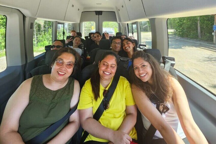 A group of people seated in a vehicle, likely a bus, with a road and greenery visible through the windows. 4
