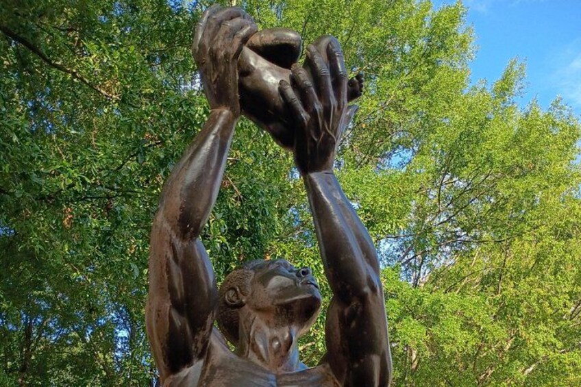En bronzestatue af en person med løftede arme foran et grønt trælandskab. 5