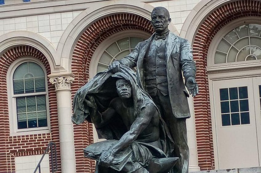 En statue af en mand stående ved siden af en knælende figur foran en bygning med buede vinduer og murstensfacade. 3