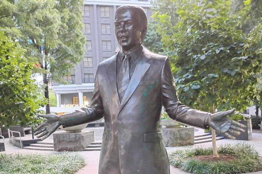 En bronzestatue af en mand i jakkesæt med armene udstrakte, placeret i et anlagt område med træer og en bygning i baggrunden. 4