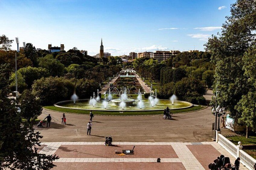 A park with a fountain, people walking, and a building in the background. 2