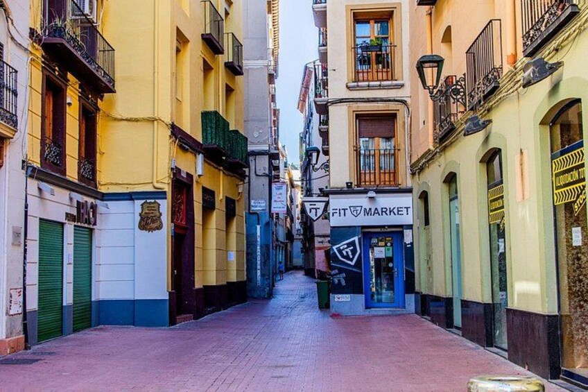 A narrow street lined with colorful buildings, a Fit Market store, and closed shop shutters. 3