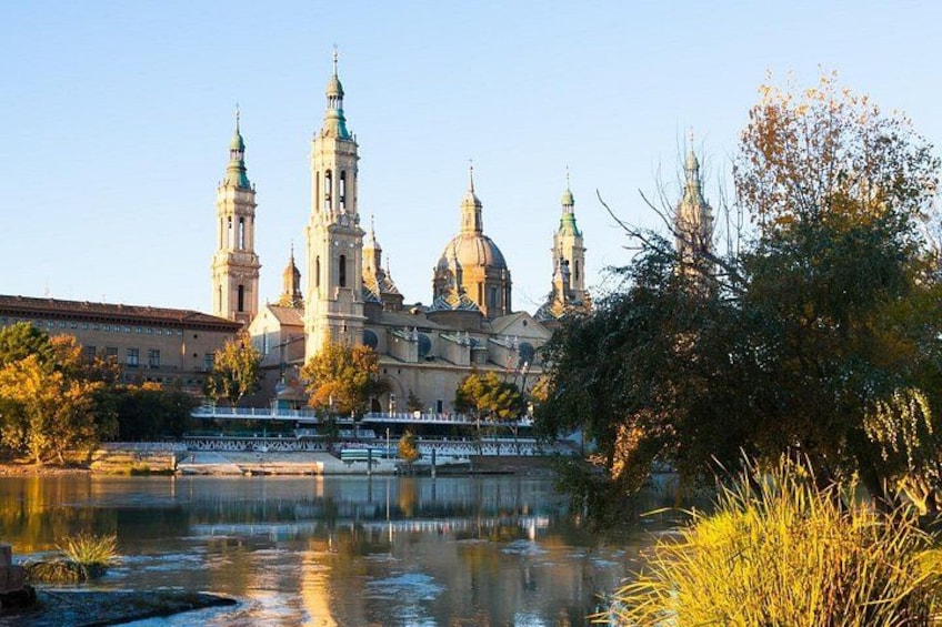 Un edificio histórico con cúpulas y torres, un río en primer plano y árboles con follaje otoñal. 1