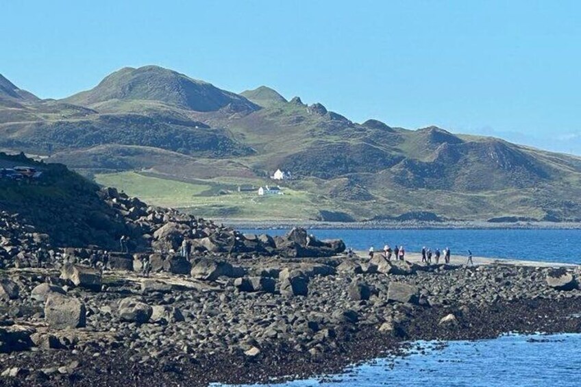 En stenet kystlinje med en hvid bygning i det fjerne, omgivet af bakker og en klar himmel. 5