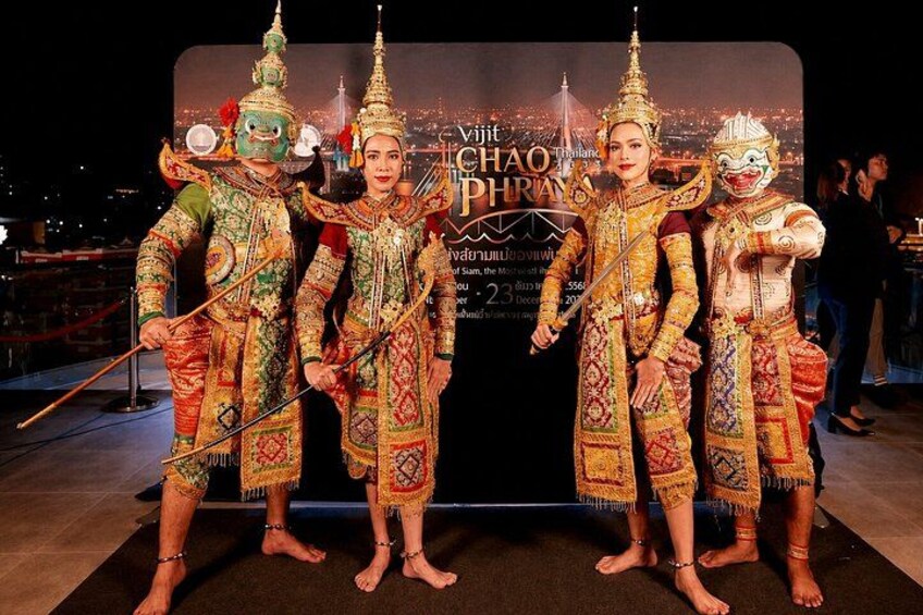 Fyra personer klädda i traditionella thailändska dräkter med utsmyckade huvudbonader och accessoarer, stående framför en bakgrund med text. 2