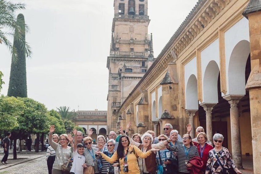 A group of people in a courtyard with a tall tower and arches in the background. 2