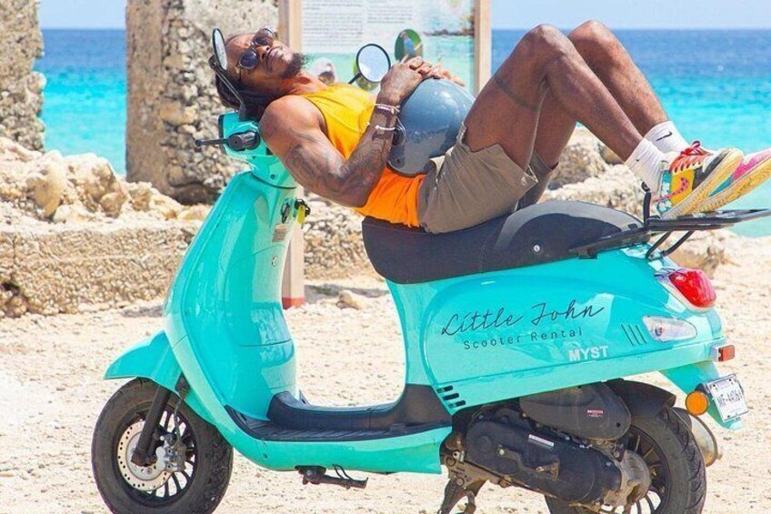 A person lying on a turquoise scooter with the text 'Little John Scooter Rental' on it, set against a beach backdrop with clear skies and the ocean in the distance. 3