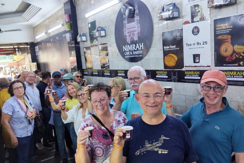 A group of people at a cafe with a sign for 'Nimrah Cafe & Bakery' and various food items displayed. 4