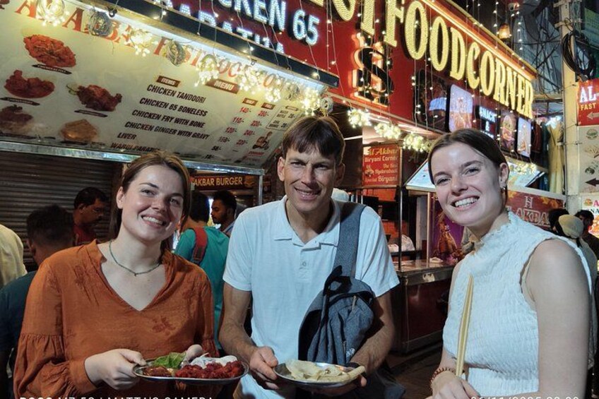 Drei Personen halten Essen an einem Essensstand, auf dessen Speisekarte verschiedene Gerichte angeboten werden. 1